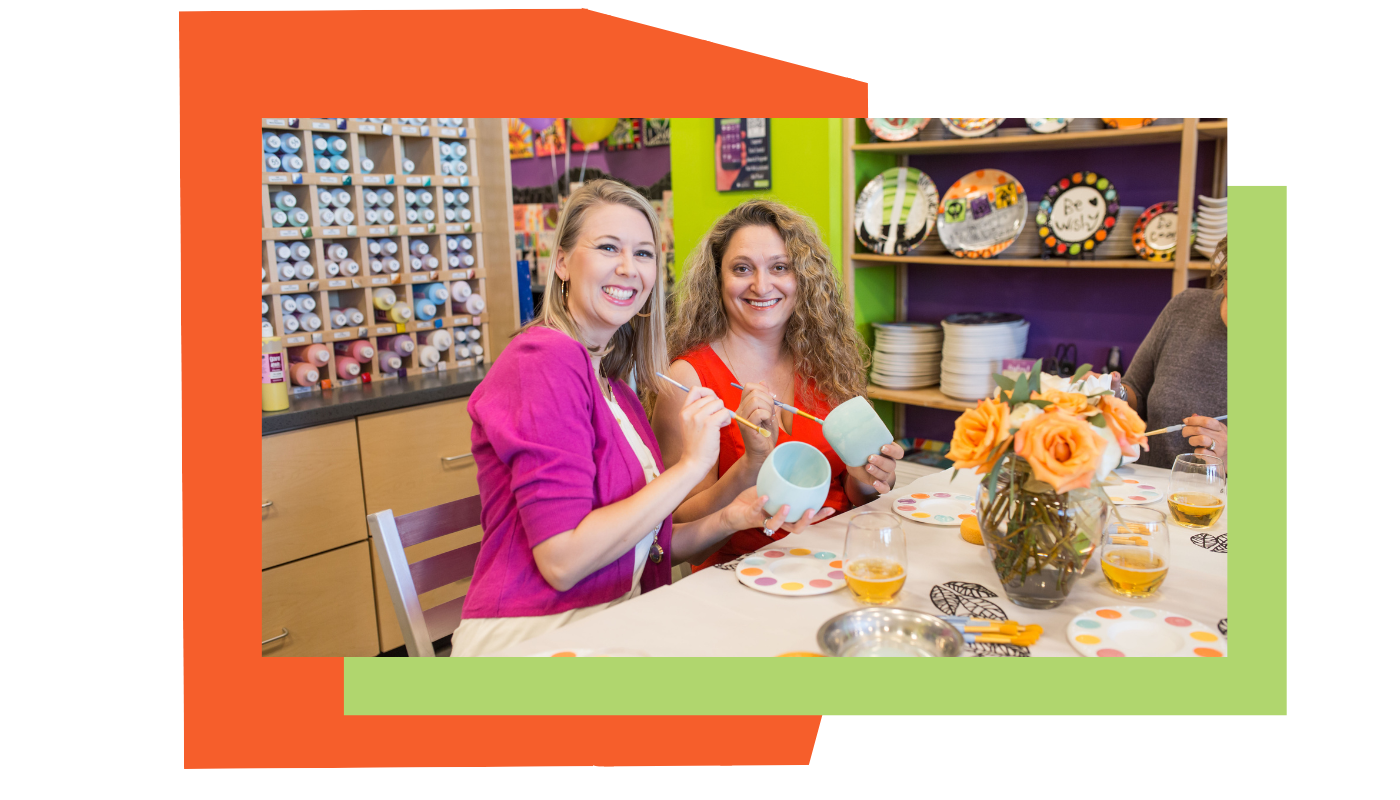 Two women at a table with ceramic items and a colorful background attending a bridal shower at As You Wish Pottery