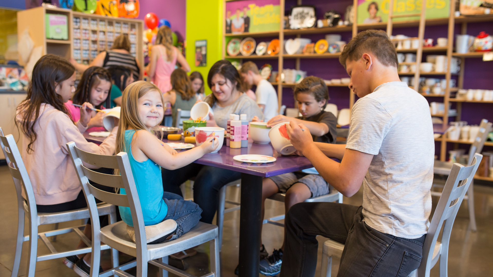 Children and young adults sitting at a table in an As You Wish Pottery studio painting ceramic bowls