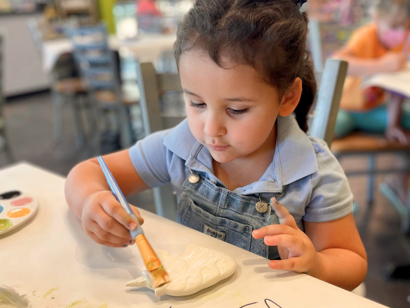 Child painting a ceramic object at a table with other children in the background at an As You Wish Pottery painting studio.