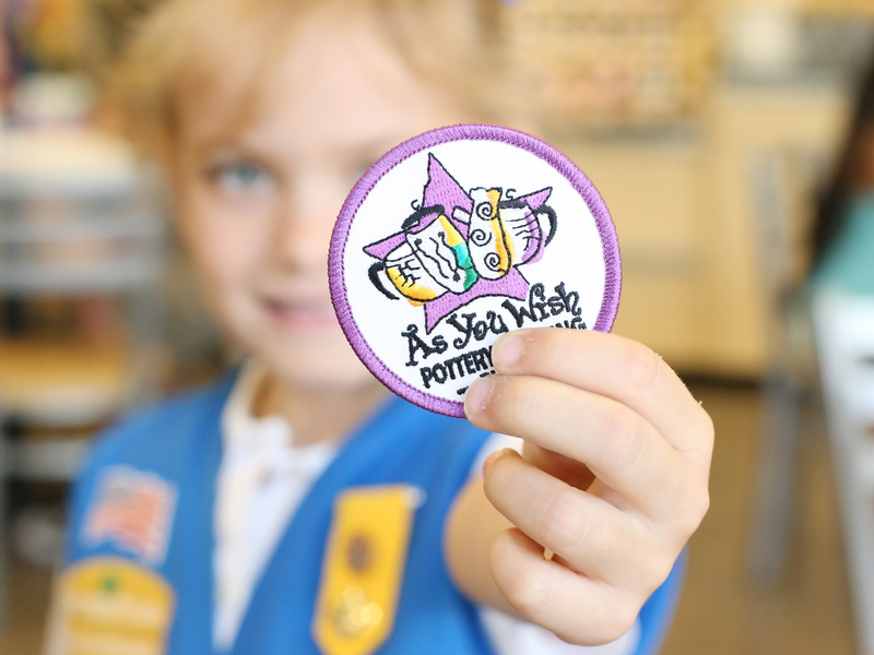 Daisy Girl Scout holding a patch with As You Wish logo, blurred background