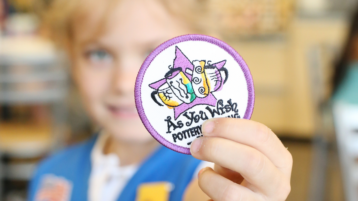 Child holding a 'As You Wish Pottery' patch with a blurred background