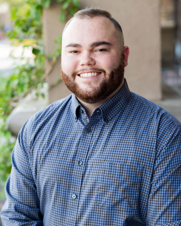 Man wearing a blue checkered shirt outdoors with plants and a building in the background